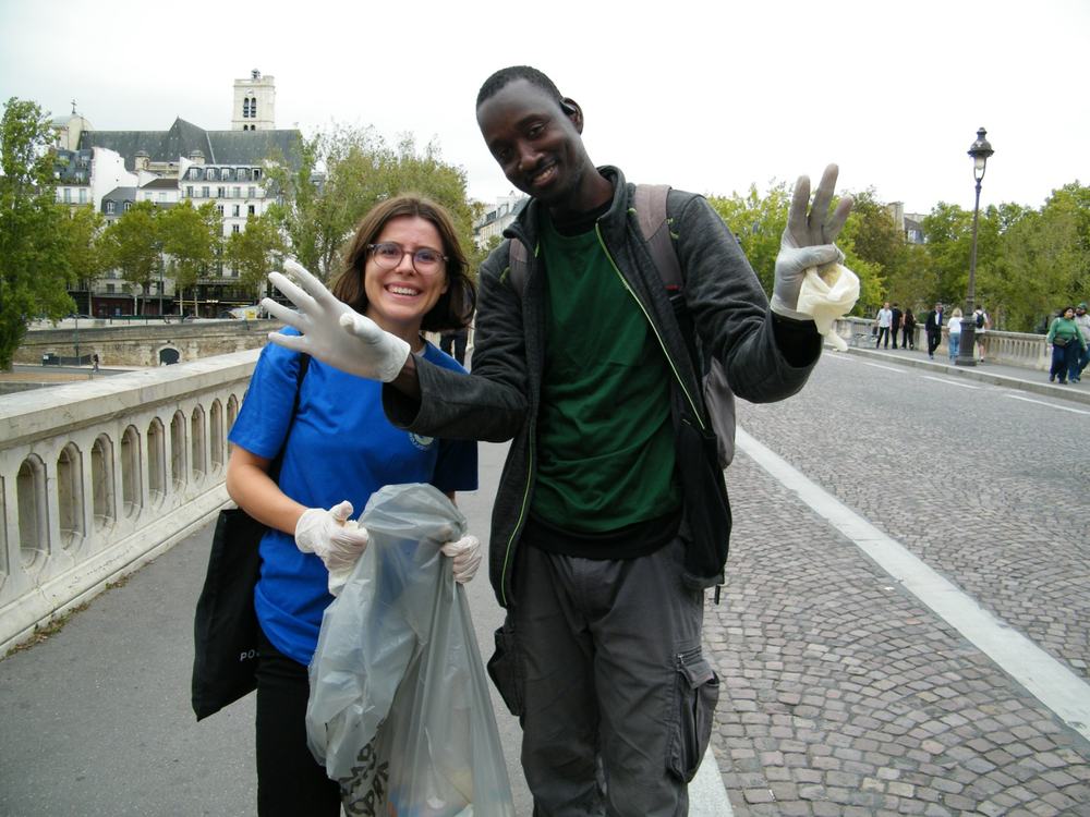 Two smiling volunteers waving on a Paris bridge, holding a bag of litter